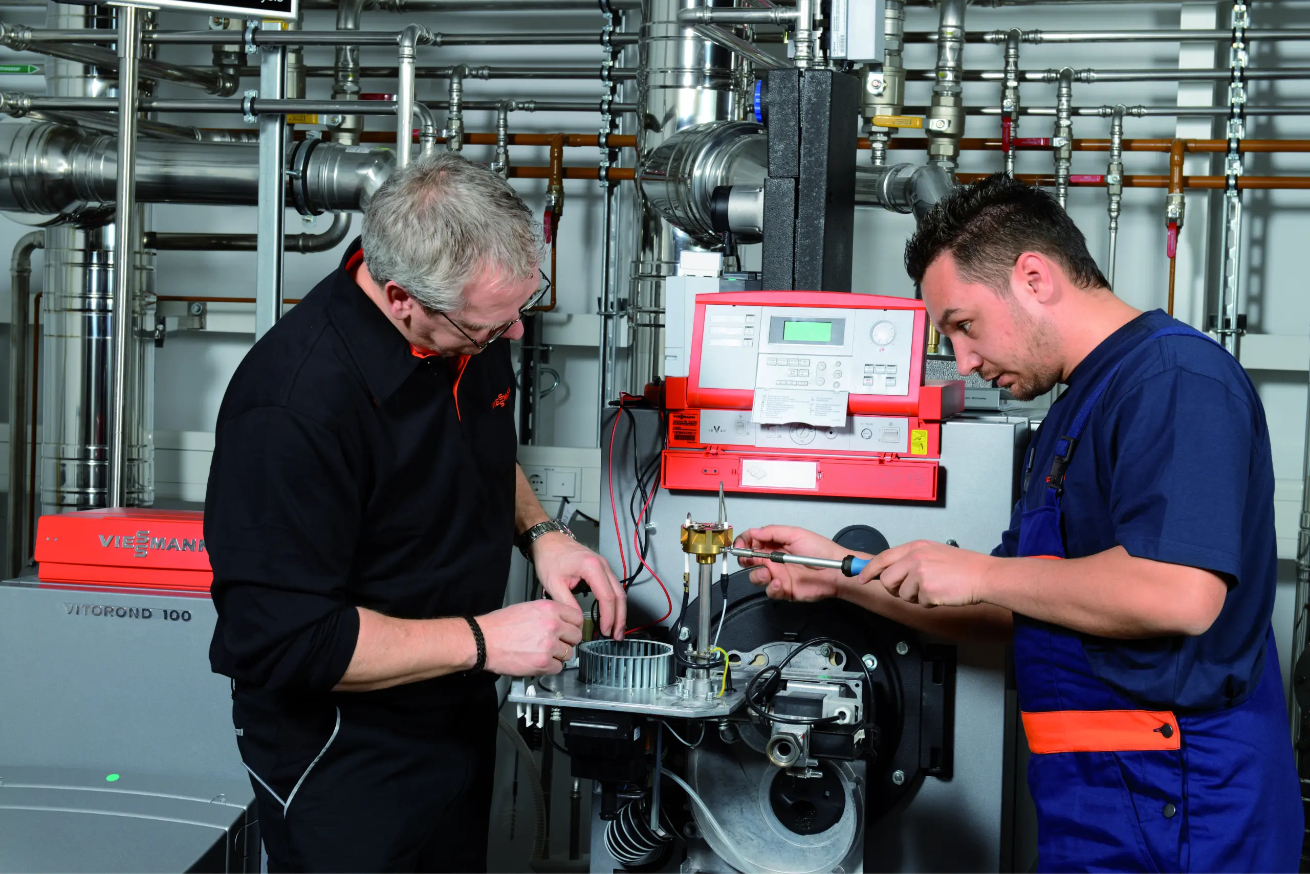 Dieses Bild zeigt zwei Techniker, die an einer Viessmann Heizanlage arbeiten. Sie sind in einem technischen Raum umgeben von Rohrleitungen und Heizungsanlagen. Der ältere Techniker auf der linken Seite trägt ein schwarzes Hemd und arbeitet an einer Komponente der Anlage, während der jüngere Techniker auf der rechten Seite, in blauer Arbeitskleidung, mit einem Schraubendreher an der Anlage arbeitet. Die Viessmann Heizanlage im Hintergrund ist gut erkennbar mit einem roten Bedienfeld und der Aufschrift "VITOROND 100". Beide Techniker sind konzentriert bei der Arbeit und scheinen eine Wartung oder Reparatur durchzuführen.