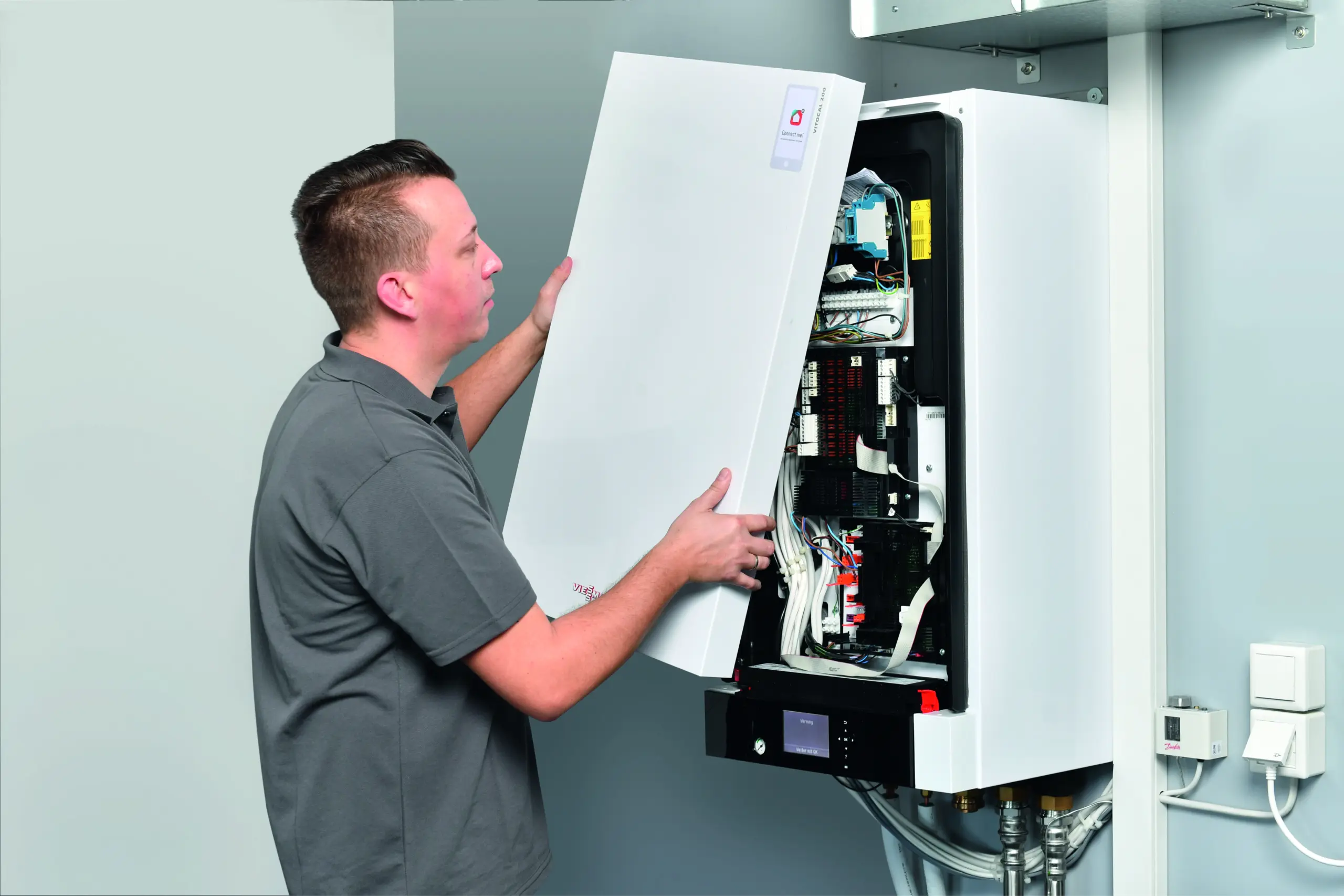 Ein Techniker überprüft ein geöffnetes elektrisches Schalttafelgehäuse von Viessmann. Er hält die Abdeckung des Gehäuses, das eine Vielzahl von Kabeln und Komponenten zeigt. Die Szene findet in einem sauberen, modernen technischen Umfeld statt. Die Installation scheint professionell und ordentlich zu sein, was auf eine sorgfältige Wartung und Inspektion hinweist.