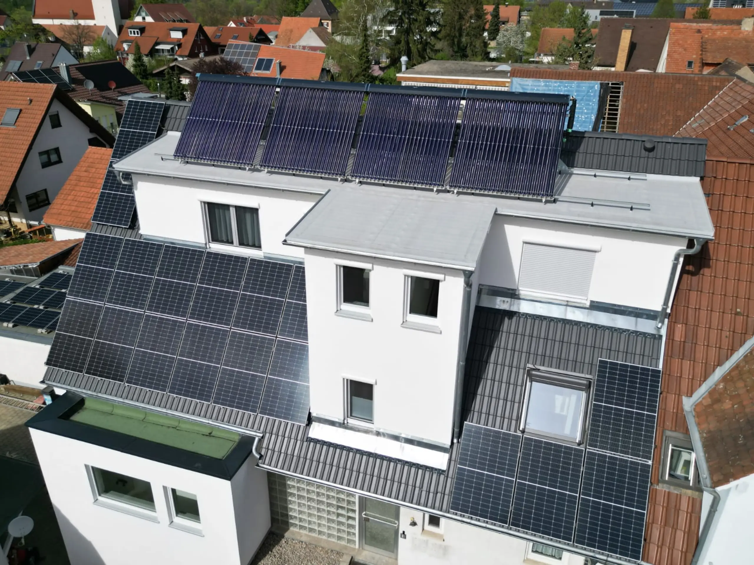 Ein Wohnhaus mit Solarmodulen und Solarthermiekollektoren auf dem Dach. Die Solarpaneele bedecken einen Großteil der Dachfläche und zeigen das Engagement des Hauses für erneuerbare Energien. Die umliegenden Gebäude in der Nachbarschaft vermitteln eine ruhige, ländliche Wohngegend.