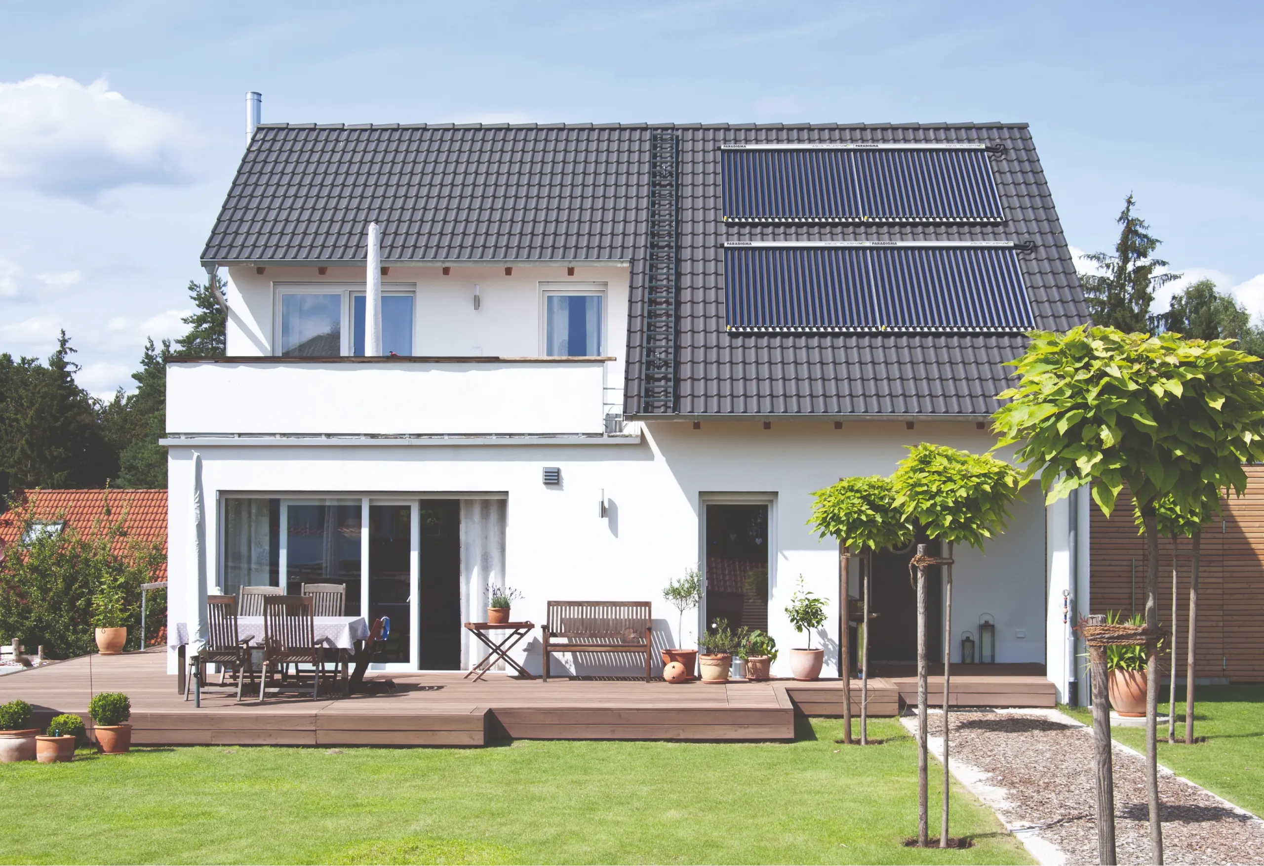 Das Bild zeigt ein modernes Einfamilienhaus mit einem gepflegten Garten. Auf dem Dach sind Solarpaneele von Paradigma installiert, die in zwei Reihen angeordnet sind und sich nahtlos in das Design des Hauses einfügen. Das Haus verfügt über eine geräumige Terrasse mit Gartenmöbeln, die zum Entspannen im Gartenbereich einlädt. Im Vordergrund stehen mehrere kleine Bäume und Pflanzen, die den Charme des gut gepflegten Gartens unterstreichen. Das Solarsystem von Paradigma exemplifiziert nachhaltige und umweltfreundliche Energielösungen.