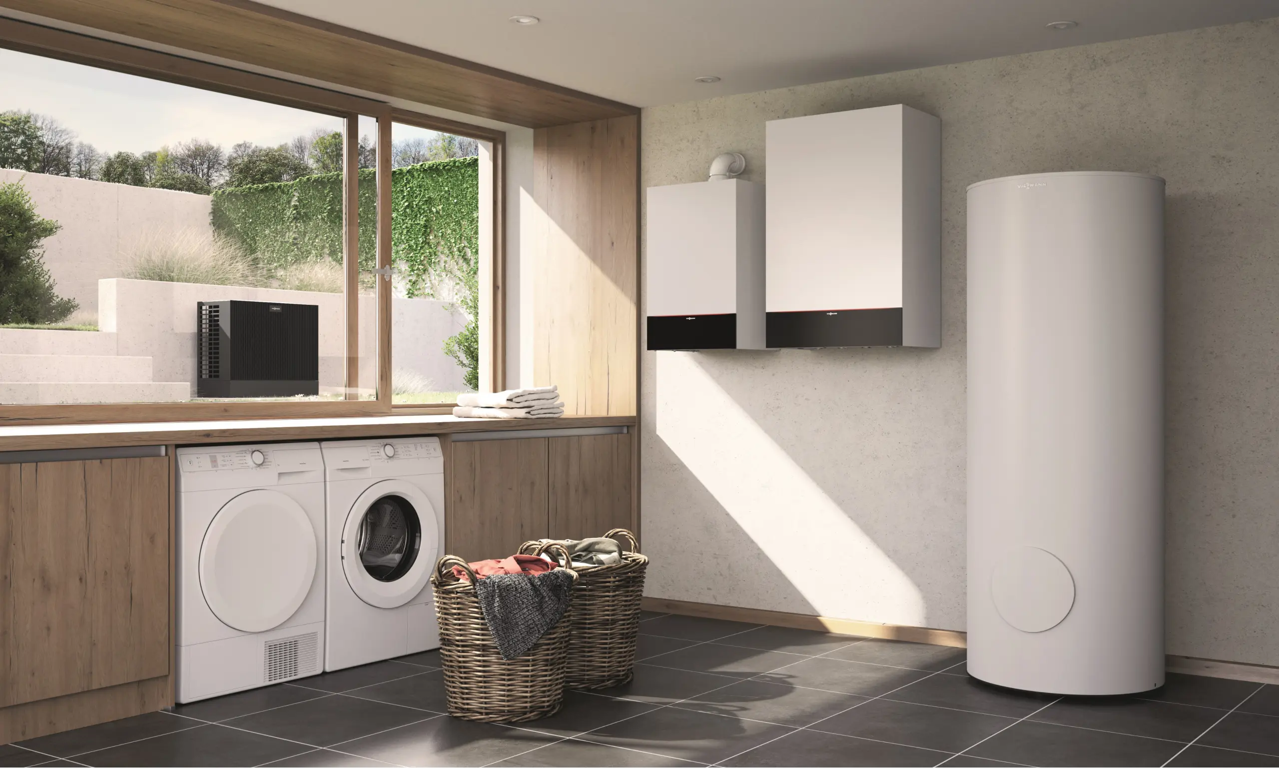 Das Bild zeigt eine moderne Waschküche mit einer großen Fensterfront, die den Raum mit natürlichem Licht durchflutet. Links unter dem Fenster befinden sich eine Waschmaschine und ein Trockner, beide in weiße Schränke integriert. Auf der rechten Seite des Bildes sind zwei Viessmann-Geräte zu sehen: die Vitocal 250-SH und der Vitodens 300-W, die nebeneinander an der Wand montiert sind. Daneben steht ein großer, zylindrischer Warmwasserspeicher. Der Raum ist mit dunklen Fliesen und Holzverkleidungen ausgestattet, die eine warme und einladende Atmosphäre schaffen. Zwei Wäschekörbe aus geflochtenem Material befinden sich auf dem Boden, bereit für die nächste Ladung Wäsche. Durch das Fenster ist ein gepflegter Garten zu sehen, was dem Raum eine frische und ruhige Note verleiht.